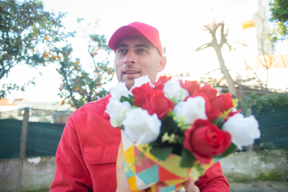 A male flower delivery courier from Biggin Hill Florist, dressed in a red uniform and matching cap, is holding a vibrant bouquet of fresh flowers close to the camera. The bouquet features a mix of red