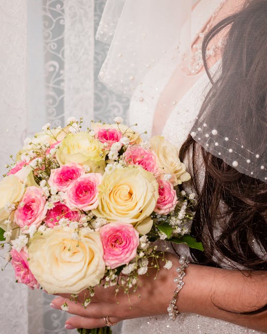 A bride holding a floral bouquet arrangement featuring large, creamy white roses and smaller pink roses, interspersed with delicate white baby's breath flowers. The bouquet is wrapped in soft white pa