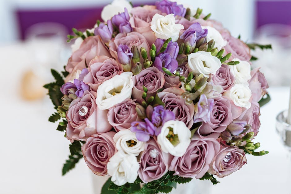 A round floral bouquet featuring soft pink roses, white lisianthus, and purple freesia arranged in a dense, harmonious cluster. The flowers rest atop a white table with subtle reflections, and the bou