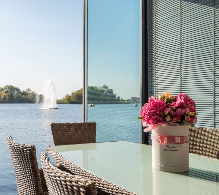 A round white floral gift box filled with an arrangement of fresh pink flowers, including hydrangeas, roses, and smaller filler blooms, is placed on a glass-topped outdoor table. The bouquet features 