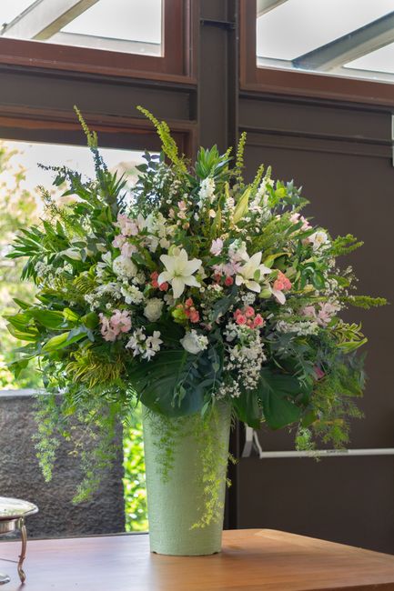 A tall, elegant floral arrangement featuring a mix of fresh flowers including white lilies, pink roses, and delicate white and pink blossoms, arranged in a lush, abundant style with greenery such as l