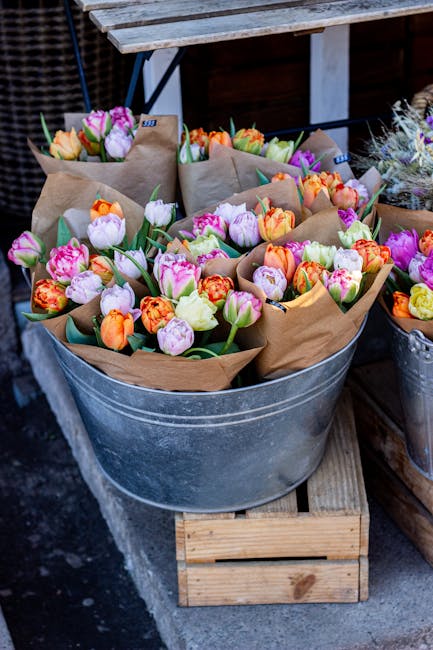 A large galvanized metal bucket filled with multiple vibrant bouquets of tulips in shades of pink, orange, purple, and yellow, each wrapped in brown kraft paper. The tulips display freshly opened peta