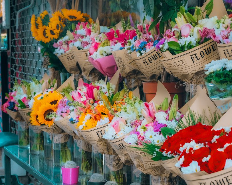 An assortment of fresh floral bouquets displayed on a glass shelf in a floristry shop. The bouquets include vibrant sunflowers, pink lilies, white roses, and red carnations, each wrapped in rustic pap