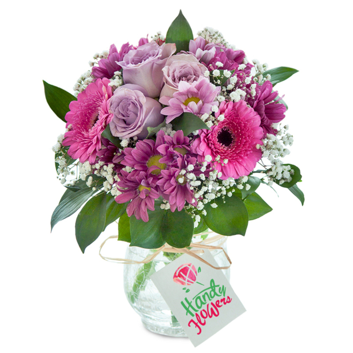 Emanating vibrant color and sophistication, this meticulously designed floral arrangement captures Biggin Hill's vibrant local character. Set against a luminous white background, the bouquet overflows with the harmonious blend of blush pinks, deep purples, and gentle lavenders. At the arrangement's heart, soft lavender roses blossom with timeless grace. Surrounding them, lively fuchsia Gerbera daisies with striking dark centers inject a burst of cheerful energy, while clusters of magenta chrysanthemums with hints of yellow-green provide subtle contrast and depth. The bouquet is lightened by generous tufts of white baby's breath, their frothy texture reminiscent of spring clouds drifting above Greenacres. Lush, dark green foliage envelops the blooms, grounding the composition in natural beauty. The bouquet is arranged in a clear, textured glass vase reminiscent of artisan craftsmanship, the surface playing with light like reflections on rain-washed pavement. Tied at the neck with natural raffia ribbon and finished with a neat white tag, the arrangement awaits a message of celebration, love, or appreciation-perfect for expressing heartfelt sentiment or marking meaningful moments throughout Biggin Hill. The artistry and freshness of this bouquet echo the care and creativity found at Biggin Hill Florist, delivering warmth with every petal.